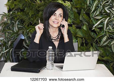 Stock Image - Professional businesswoman talking on her phone and working on her laptop computer in cafeteria; Edmonton, Alberta, Canada. Fotosearch