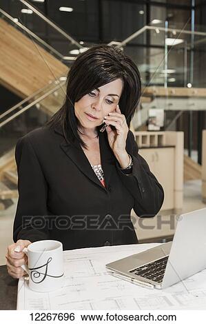 Professional businesswoman talks on phone while reviewing construction plans for office interior; Edmonton, Alberta, Canada View Large Photo Image Stock Photograph - Professional businesswoman talks on phone while reviewing construction plans for office interior; Edmonton, Alberta, Canada. Fotosearch