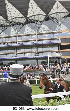 Stock Image - Rear view of a top hatted gentleman viewing horses in the Parade Ring with new Enclosure stand. Royal Ascot horse race meeting. Ascot, Berkshire, England., Ascot, Berkshire, England.. Fotosearch