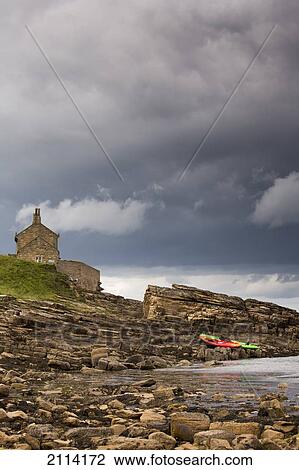 Stock Image - Red And Green Kayaks Sitting On The Rocky Shore With A House At The Water's Edge; Northumberland England. Fotosearch