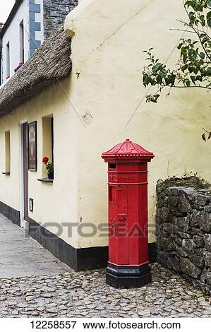Red mailbox standing on cobblestone next to stone fence with thatched roof building in background; Bunratty, County Clare, Ireland View Large Photo Image Stock Photo - Red mailbox standing on cobblestone next to stone fence with thatched roof building in background; Bunratty, County Clare, Ireland. Fotosearch