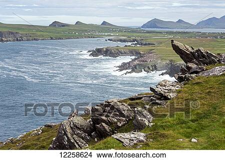 Picture - Rugged rocks in the hillside overlooking rough coastline and flat grasslands and mountains in the distance; Ballinglanna, County Cork, Ireland. Fotosearch