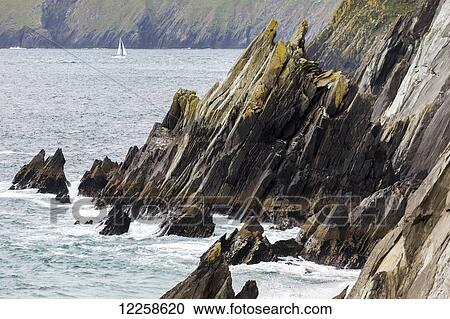 Stock Image - Rugged rocky shoreline with sailboat in the distance; Dingle, County Kerry, Ireland. Fotosearch