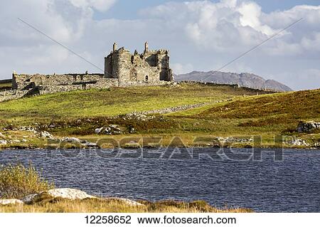 Stock Image - Ruined stone castle on rolling hill with pond in the foreground and blue sky and clouds; Ballyconneely, County Galway, Ireland. Fotosearch