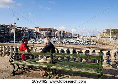 Stock Image - Senior Couple Sitting On Bench At Lekeitio Harbor, Basque Country, Spain. Fotosearch
