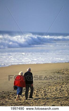 Stock Image - Senior Couple Walking On Beach View From Behind, Arm In Arm, White Shorebreak. Fotosearch