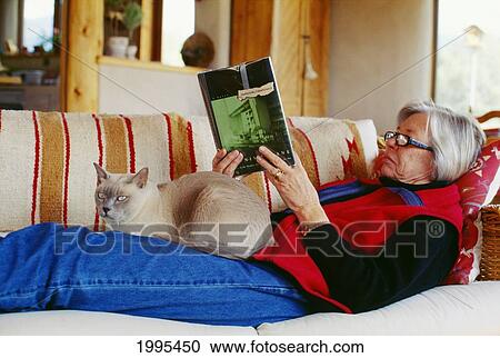 Stock Image - Senior Woman Laying On Couch Read A Book, With Cat Sitting On Lap.. Fotosearch