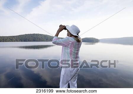 Stock Photo - Senior Woman Looking Through Her Binoculars While Standing On Her Cottage Dock In Algonquin Park; Ontario Canada. Fotosearch