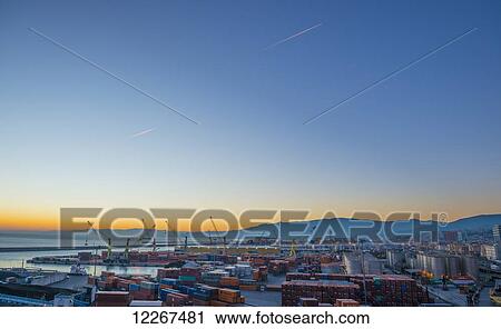 Stock Image - Shipping containers and cranes at the port at sunset with an airplane in the sky; Genoa, Liguria, Italy. Fotosearch