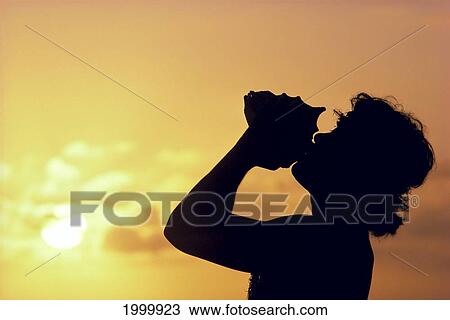 Stock Image - Side View Of Conch Shell Blower On Cliff, Silhouetted At Sunset, Pink Golden Skies. Fotosearch