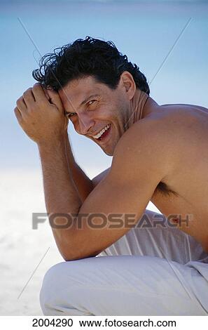 Stock Image - Side View Of Young Man Outdoor Wearing Long Pants, Smiling Head Rests In Hand Shirtless Ocean Background Blur Soft Focus. Fotosearch