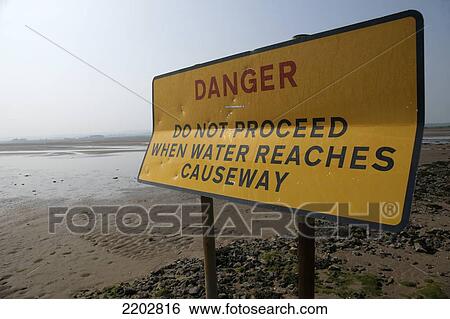 Stock Photograph - Sign warning of the danger of tidal flooding, which occurs twice a day, on the causeway leading to Lindisfarne; Northumberland, England. Fotosearch