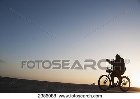 Stock Photo - Silhouette of a cyclist riding on the promenade beside the beach at sunset; California, United States of America. Fotosearch