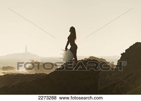 Stock Photo - Silhouette of a female surfer holding her surfboard while standing on a rock at the coast; andalusia spain. Fotosearch
