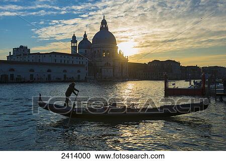Stock Image - Silhouette of a gondolier rowing his gondola with Salute church along the shoreline in the distance at sunset; Venice, Veneto, Italy. Fotosearch