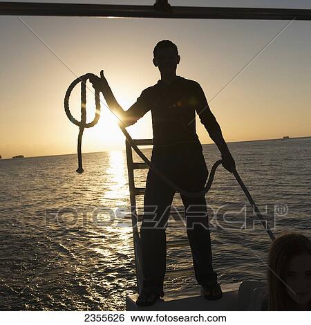 Stock Photograph - Silhouette Of A Man Standing With A Rope On The Edge Of A Boat At Sunset; Bay Islands, Honduras. Fotosearch
