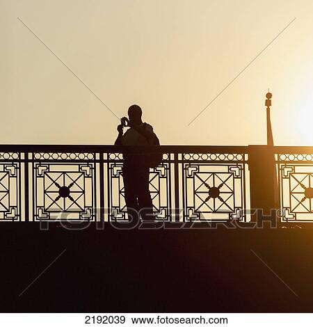 Stock Photo - Silhouette Of A Person Standing At A Railing Taking A Picture With A Camera At Sunset; Chicago Illinois United States Of America. Fotosearch