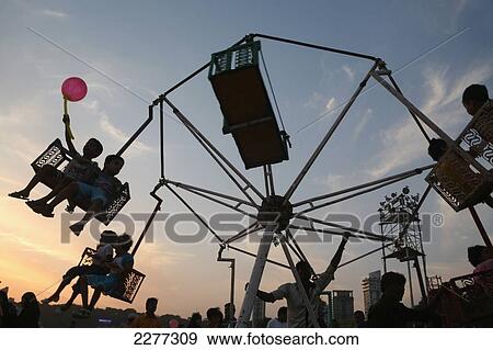 Stock Photo - Silhouette of child with balloon and children on fair ground ride at sunset at Chowpatty Beach, new development on Malabar Hill in the background; Mumbai, Maharashtra State, India. Fotosearch