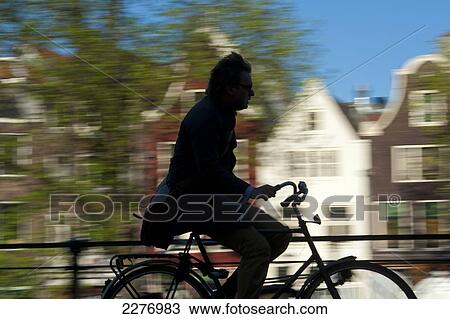 Stock Image - Silhouette of cyclist going past canal and gabled houses; Amsterdam, Holland. Fotosearch