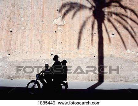 Stock Photo - Silhouette Of Father And Two Sons On Moped Going Past Shadow Of Date Palm On City Walls. Fotosearch