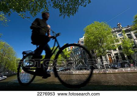 Stock Image - Silhouette of man cycling past canal; Amsterdam, Holland. Fotosearch