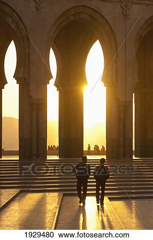 Stock Image - Silhouette of two people walking towards steps and archways with the sun glowing; Casablanca, Morocco. Fotosearch