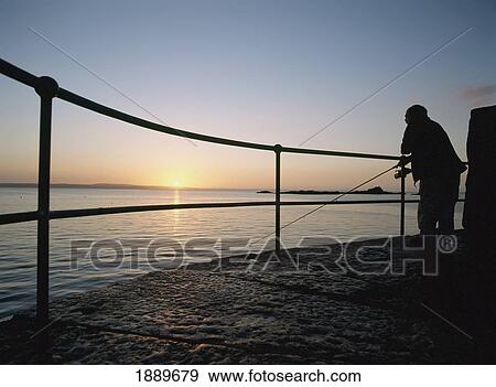 Stock Photo - Silhouetted Man Fishing Off The End Of Mousehole Harbor At Dawn. Fotosearch