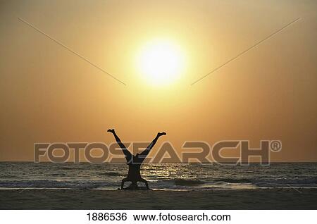 Stock Photograph - Silhouettes Of Man Doing Yoga By Ocean On The Beach At Sunset. Fotosearch