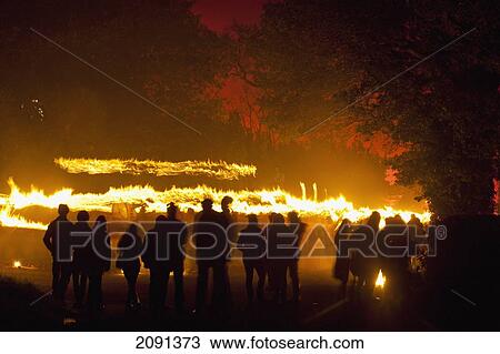 Stock Image - Silhouttes Of People Watching Procession Of Bonfire Societies With Burning Torches Coming Down Country Lane With Sky Lit By Red Flares On Bonfire Night, Barcombe, East Sussex, Uk. Fotosearch