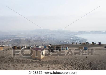 Stock Image - Small shed scattered on a sandy landscape with haze and a view of a lake; Ancon, Peru. Fotosearch