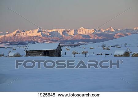Stock Photography - Snow Covered Log Cabin And Hay Bales In A Field With Illuminated Snow Covered Mountains; Okotoks, Alberta, Canada. Fotosearch