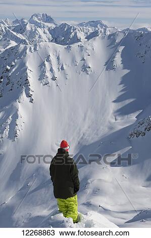 Stock Image - Snowboarder looking over the edge of a mountain with scenic snowcovered mountains in the background, Methven, New Zealand. Fotosearch