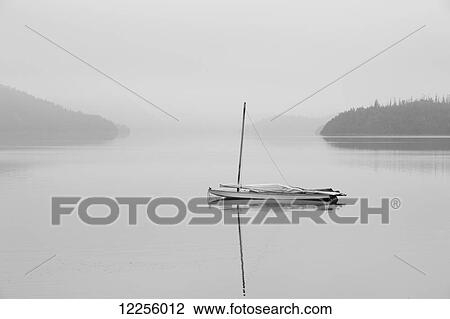 Stock Image - Solitary sailboat reflected in a misty calm lake; Ontario, Canada. Fotosearch