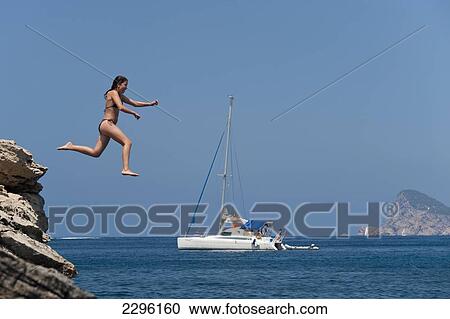 Stock Image - Spain, Girl jumping off small cliff on La Basse beach; Ibiza. Fotosearch