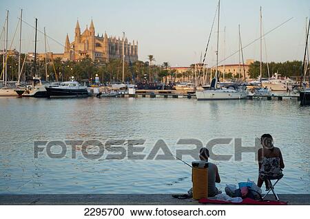 Stock Image - Spain, Majorca, Couple fishing in harbor in front of cathedral at dusk; Palma. Fotosearch