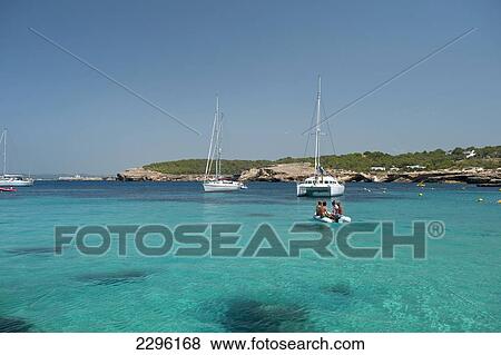 Stock Photo - Spain, People in small boat going out to their yacht off La Basse beach; Ibiza. Fotosearch