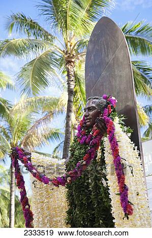 Stock Photo - Statue of a man covered in floral leis standing against a surfboard; Honolulu, Oahu, Hawaii, United States of America. Fotosearch