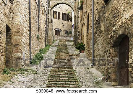 Stock Photo - Stone buildings and cobblestone walkway under an arch; Spello, Umbria, Italy. Fotosearch