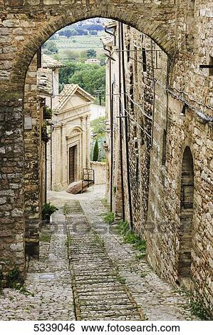 Stock Photograph - Stone buildings and cobblestone walkway under an arch; Spello, Umbria, Italy. Fotosearch