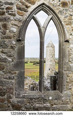 Stock Image - Stone church wall with arched window looking out at stone round tower; County Offaly, Ireland. Fotosearch