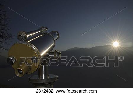 Stock Image - Sun Rays Shine Over The Mountains In The Distance With Binoculars In The Foreground; Ascona, Ticino, Switzerland. Fotosearch
