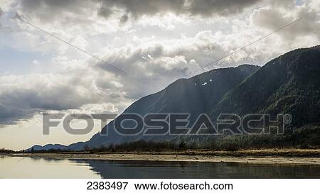 Stock Photo - Sun through clouds and shoreline of coast mountains; Coquitlam, British Columbia, Canada. Fotosearch