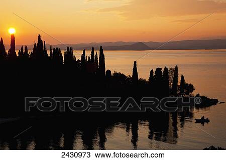 Stock Image - Sunset glowing orange over Lake Garda with a silhouette of the shoreline; Tirol, Italy. Fotosearch