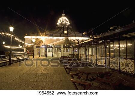 Stock Image - Tables And Chairs In A Walkway And A Building At The Pier Illuminated At Night; Eastbourne Sussex England. Fotosearch
