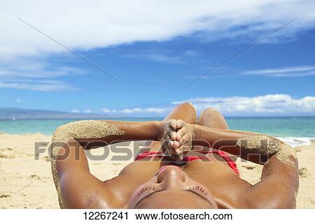 Stock Image - Tanned woman in red bikini laying on the sand at water's edge; Kauai, Hawaii, United States of America. Fotosearch