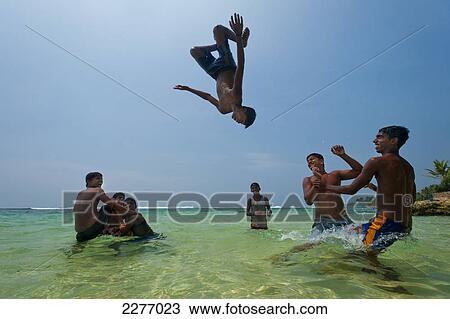 Stock Image - Teenage boys throwing each other in the air doing backflips in shallow water, Thalpe, near Unawatuna; Sri Lanka. Fotosearch