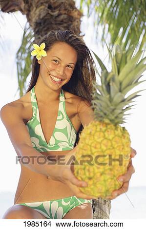 Picture - Teenage Girl Wearing A Bikini Holding Out A Pineapple, Flower In Her Hair, Palm Tree In Background.. Fotosearch