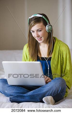 Picture - Teenage Girl With Headphones Sitting On Bed Using Laptop, Bradford, Ontario. Fotosearch