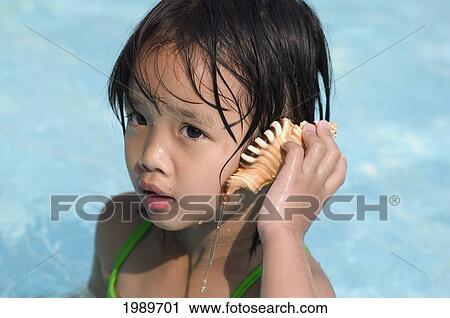Stock Image - Thailand, Nong Bai Lumphu, Five Year Old Asian Girl In Pool Listens To A Seashell.. Fotosearch
