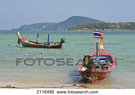 Stock Image - Thailand, Phuket, Rawaii Beach, Longtail boats along the shoreline, Land in distance.. Fotosearch